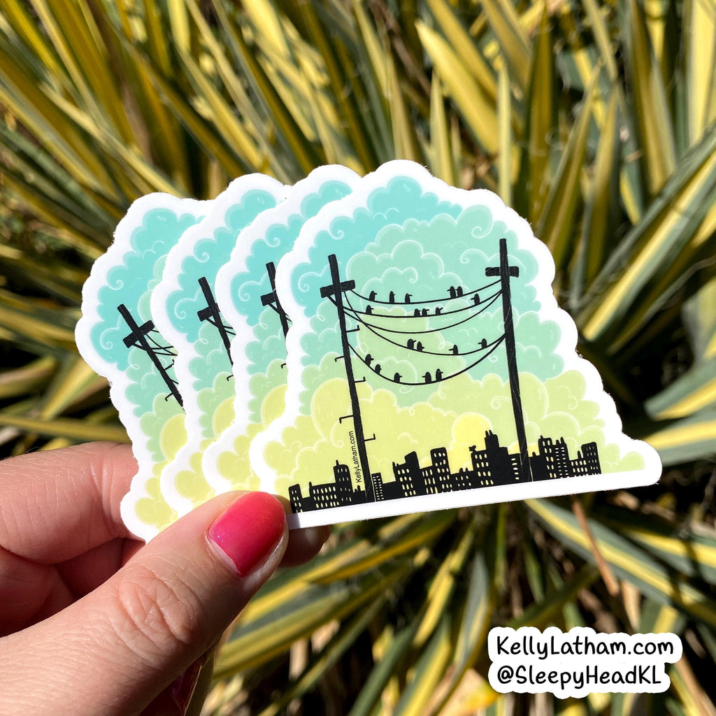 A hand with pink nail polish holds a stack of four die-cut stickers featuring a black silhouette of utility poles, wires, and a city skyline against a light blue-to-yellow gradient sky, with a blurred green and yellow plant background. A watermark in the bottom right corner reads 'KellyLatham.