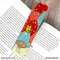 A decorative bookmark shaped like a mushroom forest, featuring red and blue mushrooms with a green frog character, placed on an open book over a wooden surface.