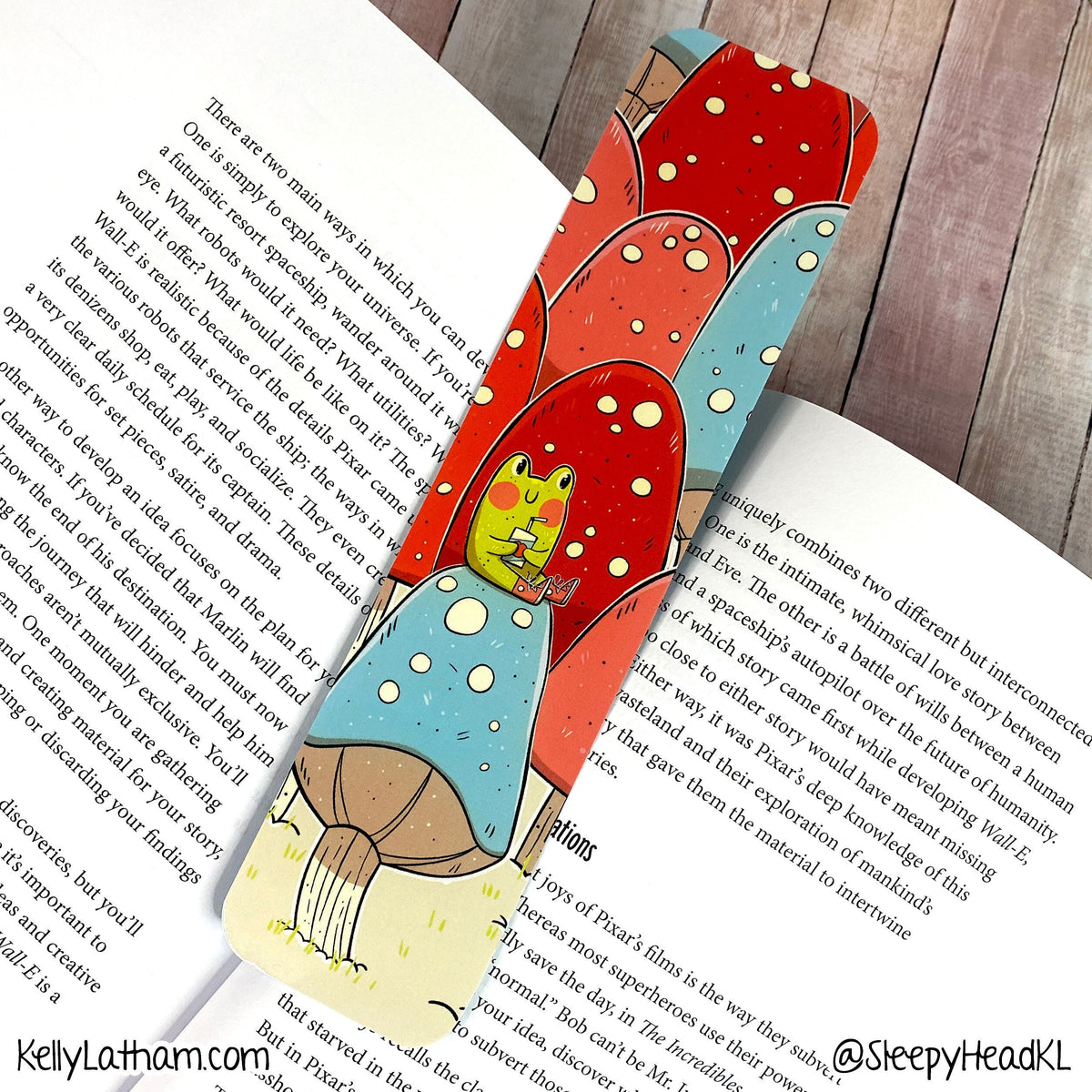 A decorative bookmark shaped like a mushroom forest, featuring red and blue mushrooms with a green frog character, placed on an open book over a wooden surface.