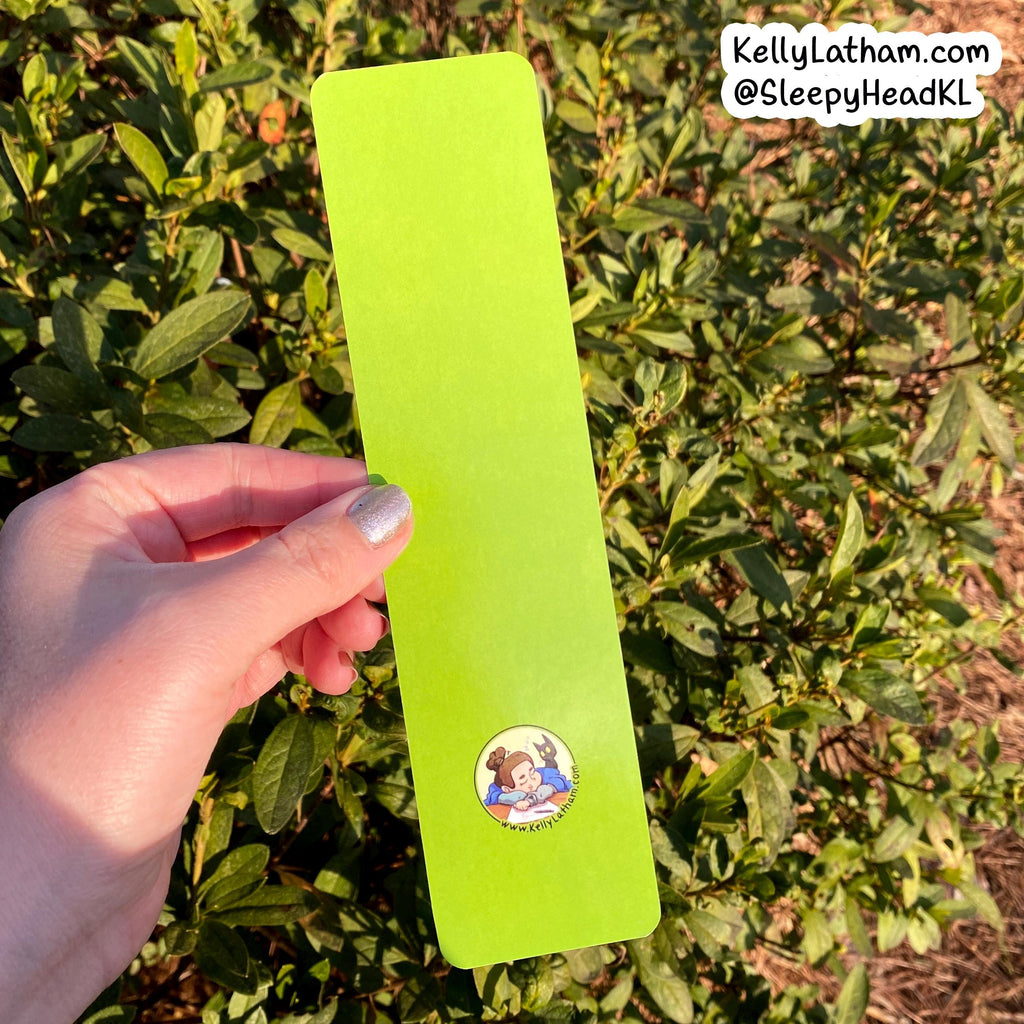 A hand holds a bright lime green rectangular card or bookmark outdoors in front of green bushes. The card has a small circular cartoon illustration near the bottom and a white text watermark in the top right corner reading 'KellyLatham.