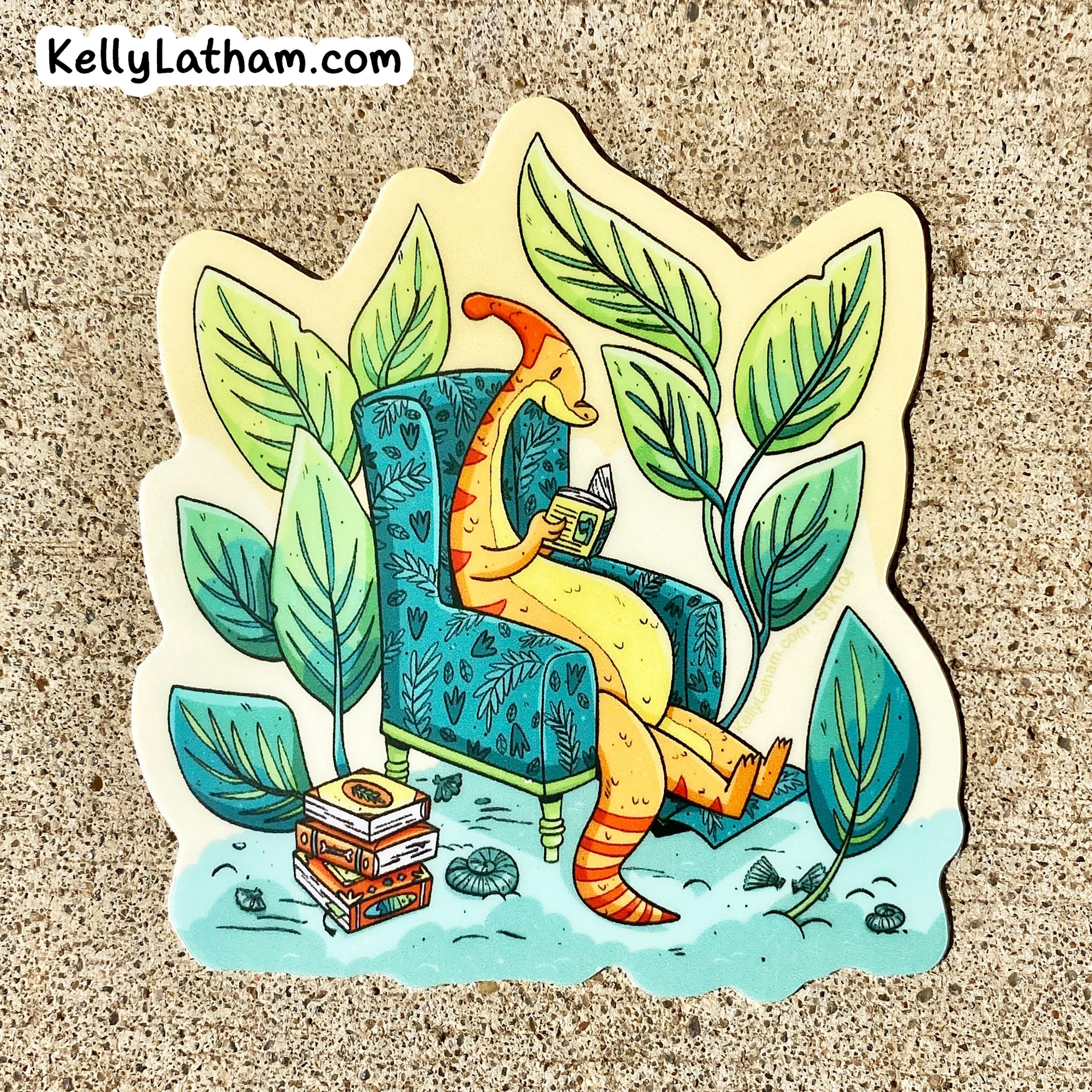 A die-cut sticker featuring an illustration of a yellow-orange dinosaur sitting in a blue patterned armchair and reading a book, surrounded by large green leaves, placed on a textured tan surface. The brand name KellyLatham.