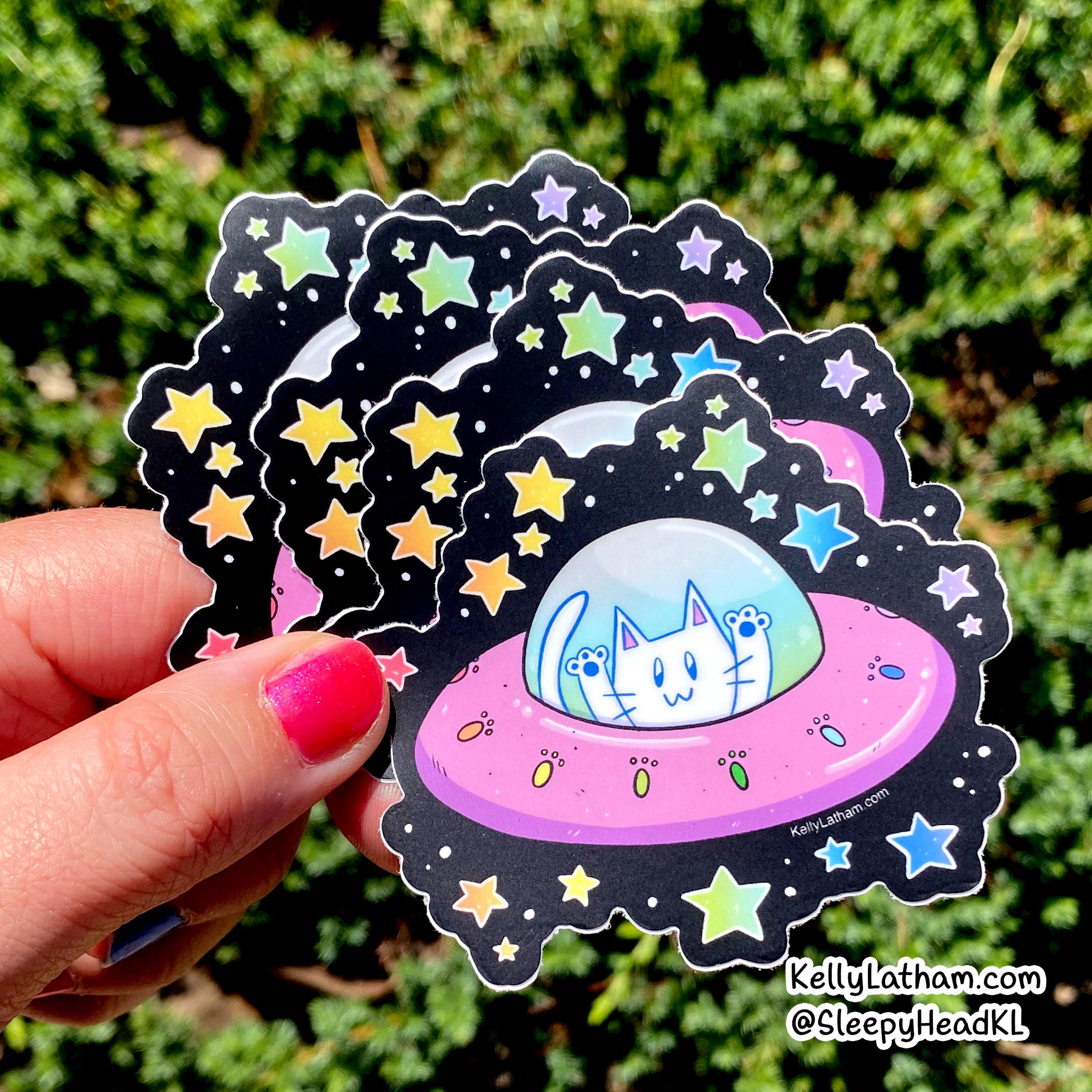 A hand with pink nail polish holds a stack of die-cut stickers featuring a cute cartoon white cat in a pink UFO surrounded by colorful stars on a black space background, with a green outdoor hedge behind it.