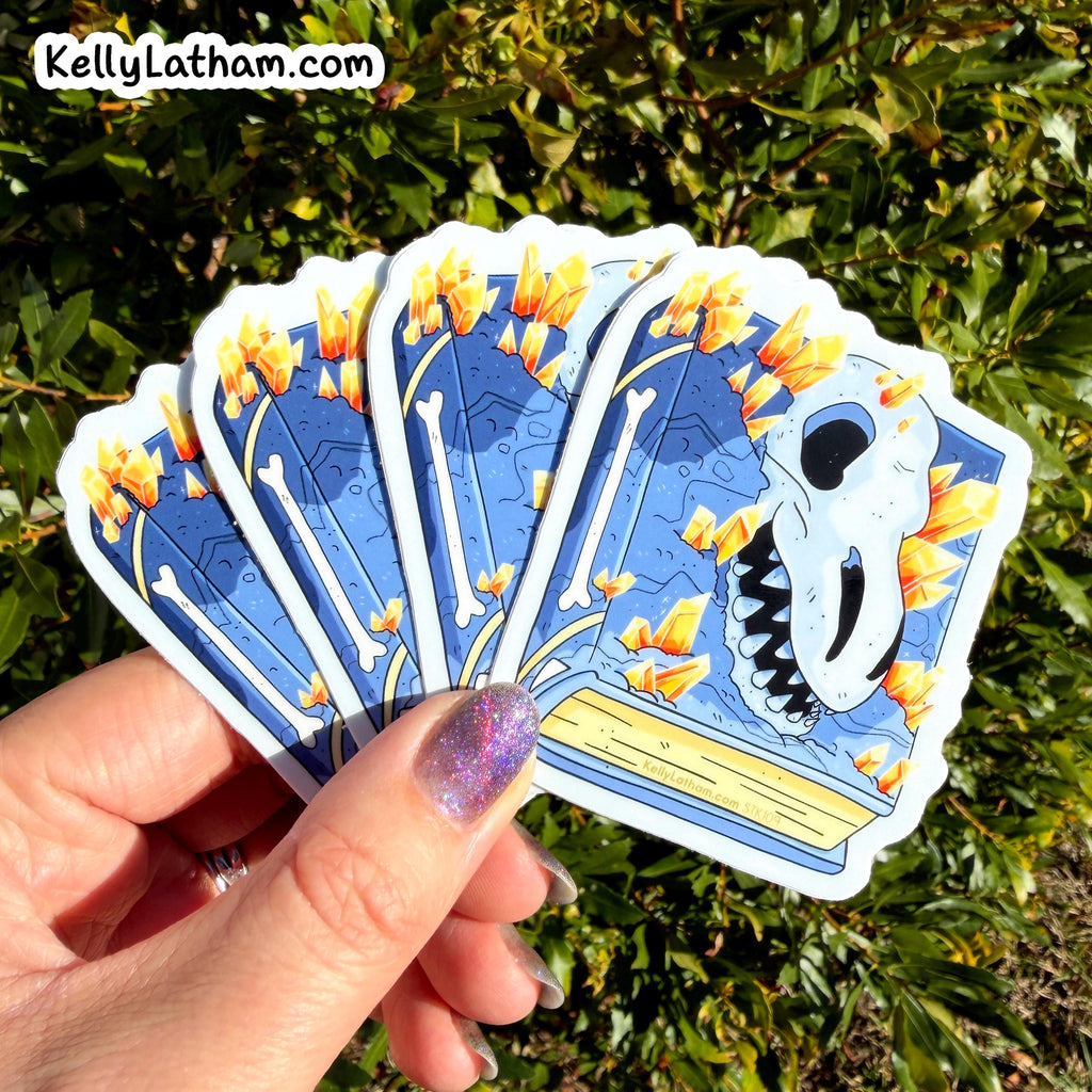 A hand with glittery purple nail polish holds a fan of five die-cut stickers featuring a blue background, white bones, and a white skull with orange flowers. The background is green foliage, and the text 'KellyLatham.