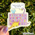 A die-cut sticker featuring a cartoon library scene with dinosaurs and colorful books, held by a hand with glittery nail polish in front of green foliage. The sticker has a speech bubble that reads 'History books are my favorite.