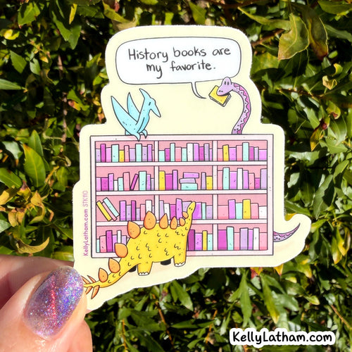 A die-cut sticker featuring a cartoon library scene with dinosaurs and colorful books, held by a hand with glittery nail polish in front of green foliage. The sticker has a speech bubble that reads 'History books are my favorite.