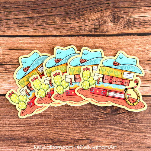 Four die-cut stickers featuring a colorful western-themed illustration with a blue cowboy hat, cactus, desert landscape, and a gold lasso, arranged on a brown wooden surface.