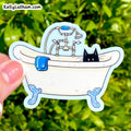 A die-cut sticker held by a hand against a blurred green outdoor background. The sticker shows a cartoon white bathtub with blue claw feet, a blue towel, a shower fixture, and a black cat peeking out.