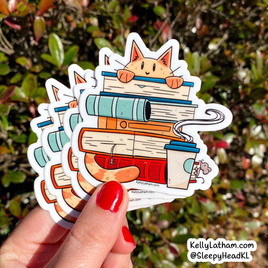 A hand with red nail polish holds a fanned-out stack of die-cut stickers featuring a cartoon cat peeking over a stack of colorful books, with a coffee cup and steam detail, set against a blurred green and red outdoor background. A watermark in the bottom right corner reads 'KellyLatham.