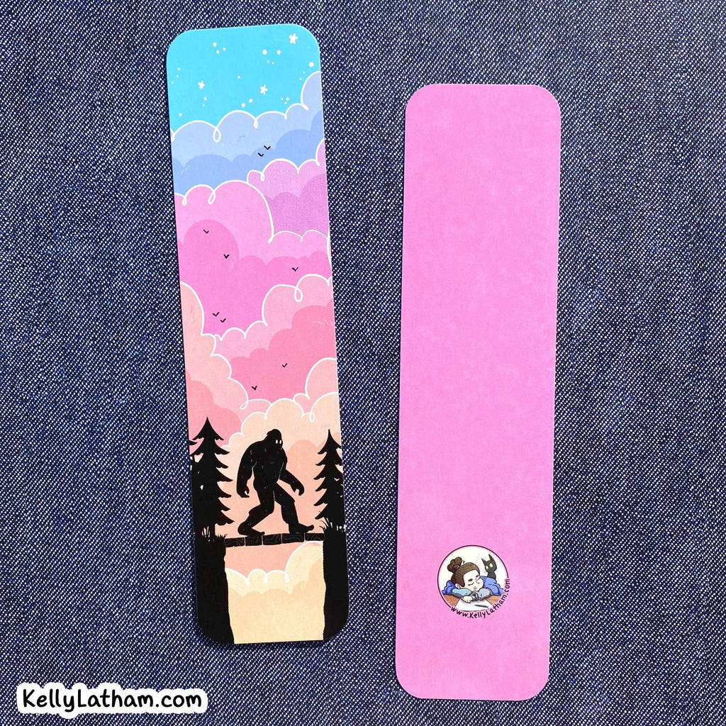 Two rectangular bookmarks laid flat on a blue denim background. The left bookmark shows a colorful illustrated sky with blue, pink, and lavender clouds, stars, birds, and a black silhouette of a Bigfoot figure above pine trees.