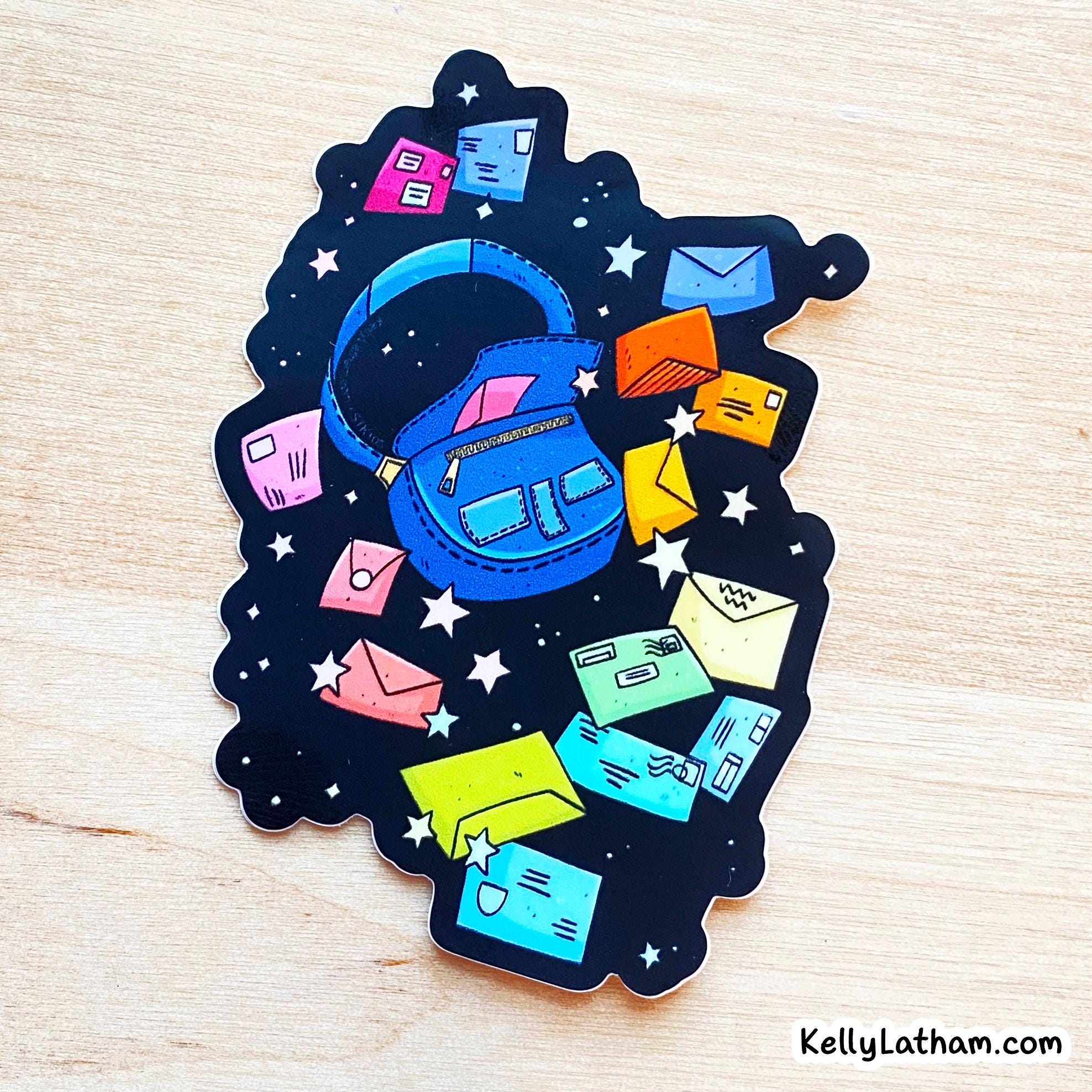 A die-cut sticker featuring a whimsical illustration of a blue backpack surrounded by colorful envelopes and cards on a black starry background, displayed on a light wood surface.