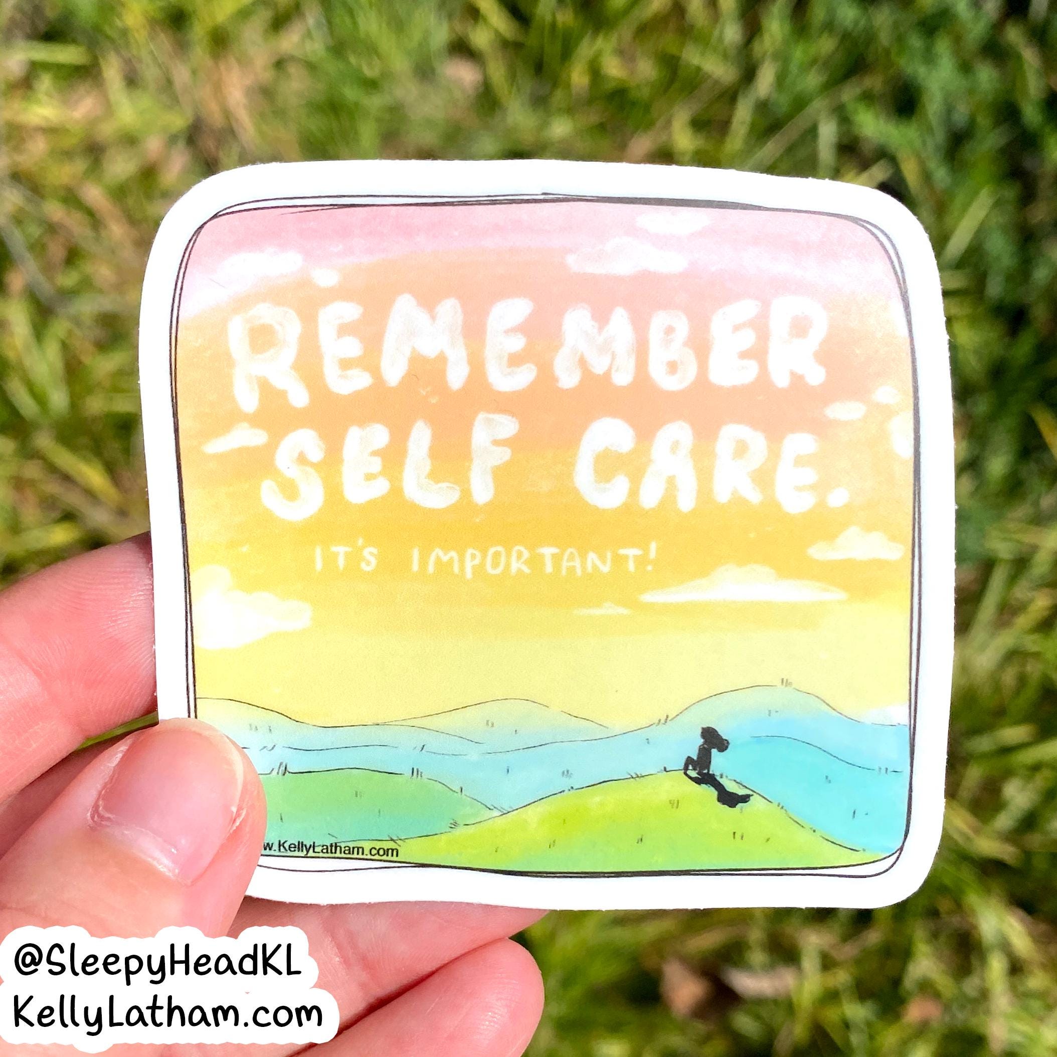 A hand holds a die-cut sticker outdoors on a grassy background. The sticker features a watercolor-style landscape with the text 'REMEMBER SELF CARE IT'S IMPORTANT!' in white lettering over a pink, yellow, and blue sky and green hills.