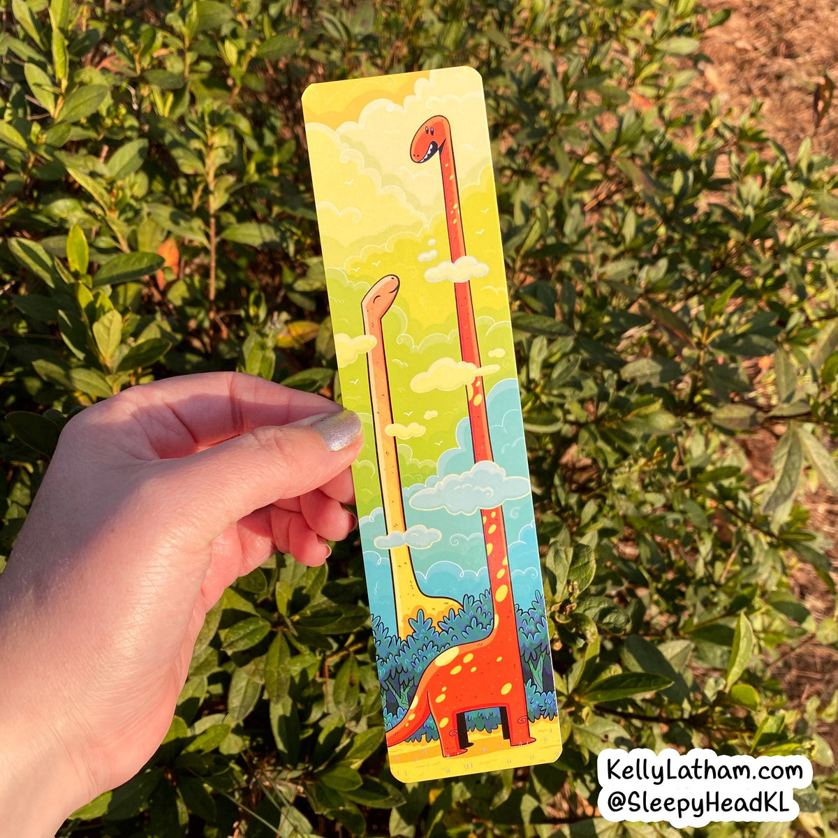 A hand holds a colorful illustrated bookmark featuring two long-neck dinosaurs under a yellow sky with clouds, set against a background of green outdoor foliage.