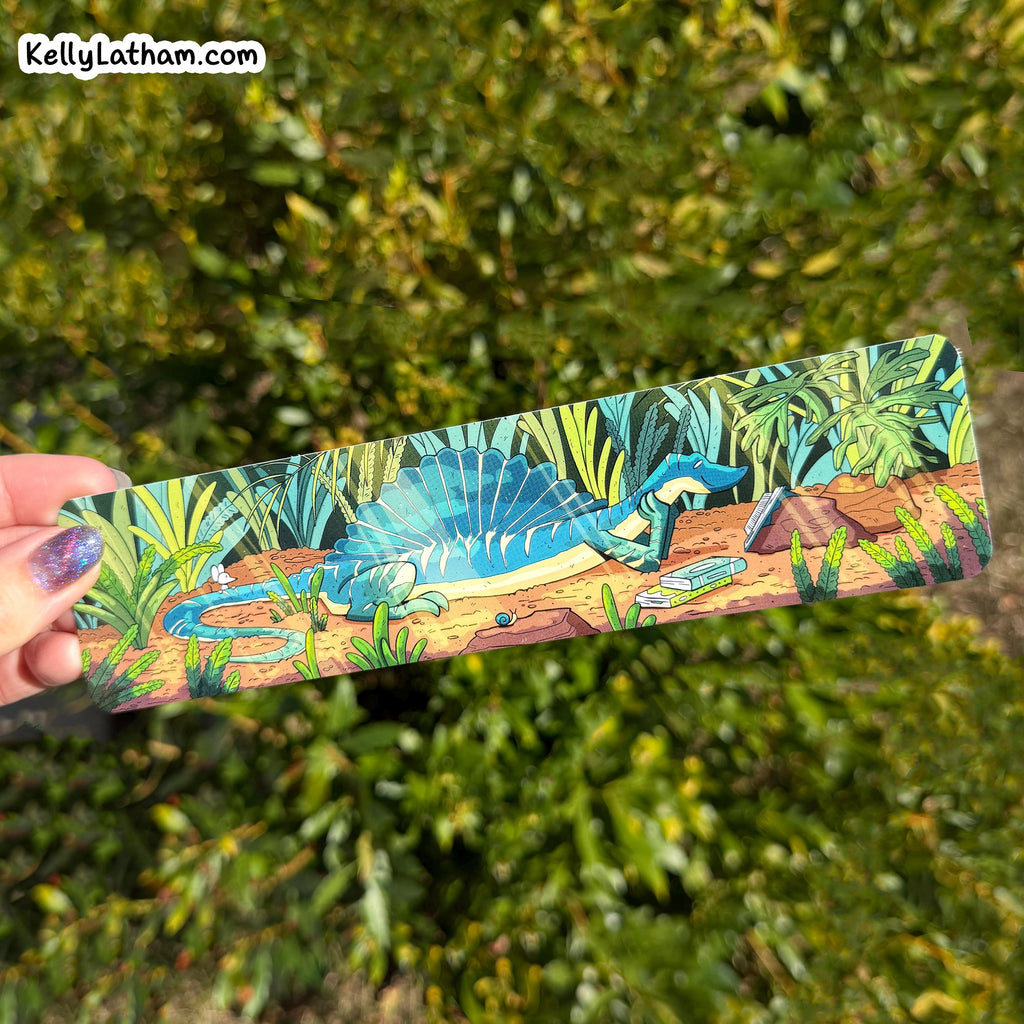A hand holds a long rectangular printed item featuring an illustrated blue spadeosaur dinosaur in a jungle scene, with a blurred green outdoor background. A small 'KellyLatham.
