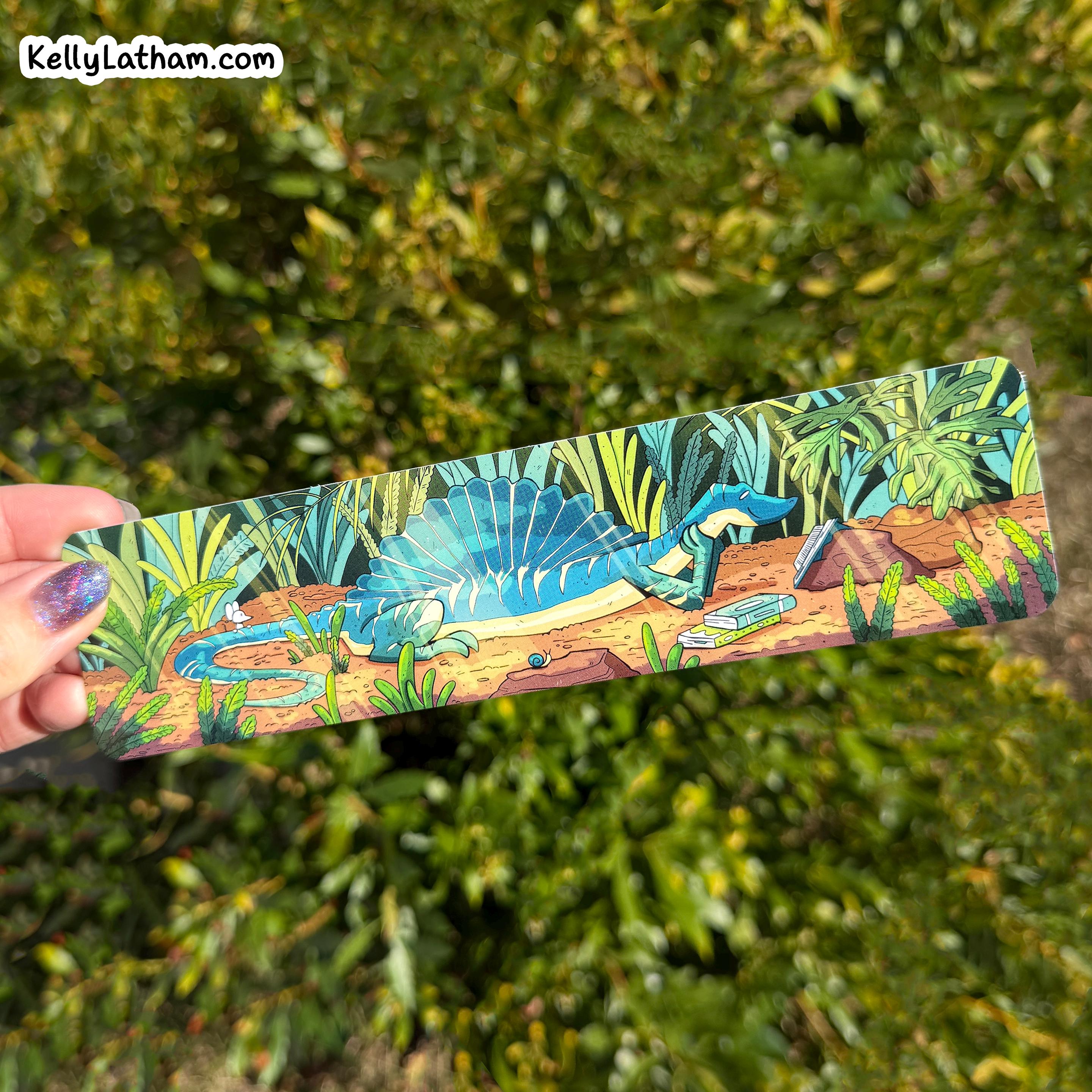 A hand holds a long rectangular printed item featuring an illustrated blue spadeosaur dinosaur in a jungle scene, with a blurred green outdoor background. A small 'KellyLatham.