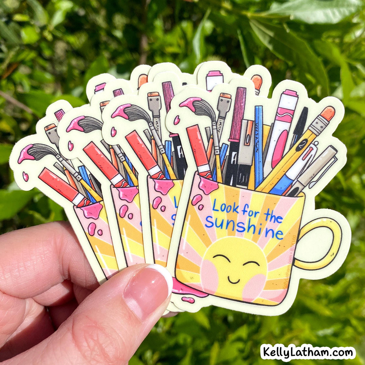 A hand holds a fan of colorful die-cut stickers featuring paintbrushes and art supplies inside a yellow mug with the text 'Look for the sunshine' against a blurred green outdoor background.