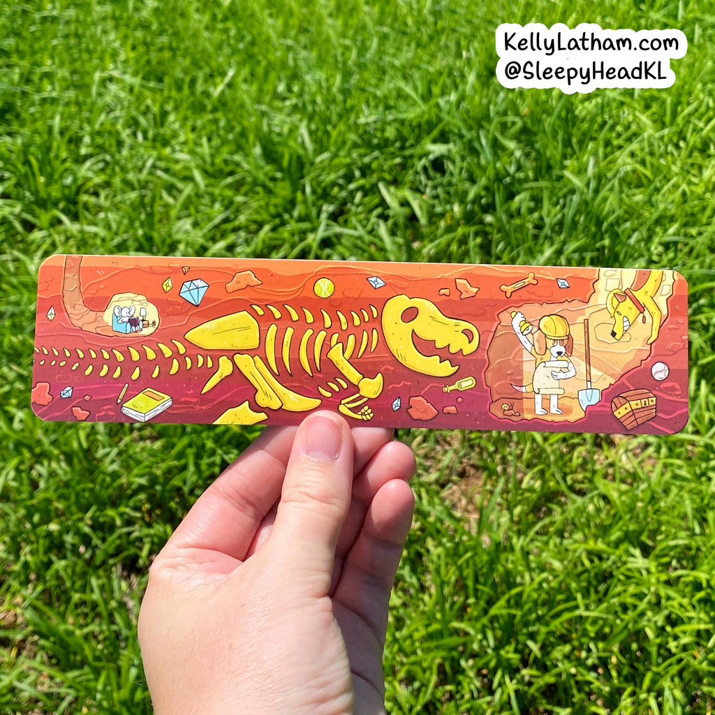A hand holds a rectangular printed card or sticker featuring a colorful dinosaur skeleton and fossil-themed illustration, photographed outdoors against a green grass background. A small watermark in the top right reads KellyLatham.