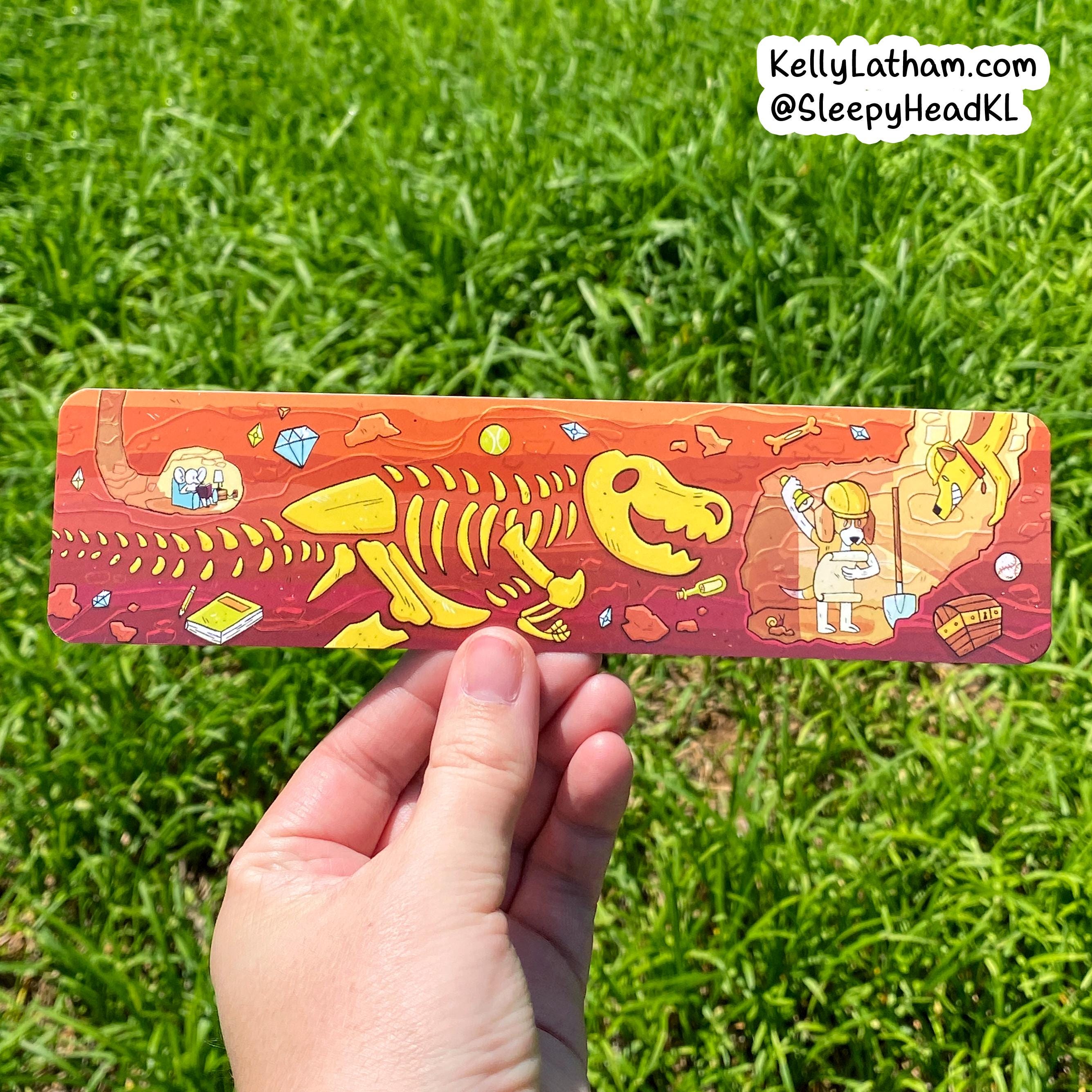 A hand holds a rectangular printed card or sticker featuring a colorful dinosaur skeleton and fossil-themed illustration, photographed outdoors against a green grass background. A small watermark in the top right reads KellyLatham.