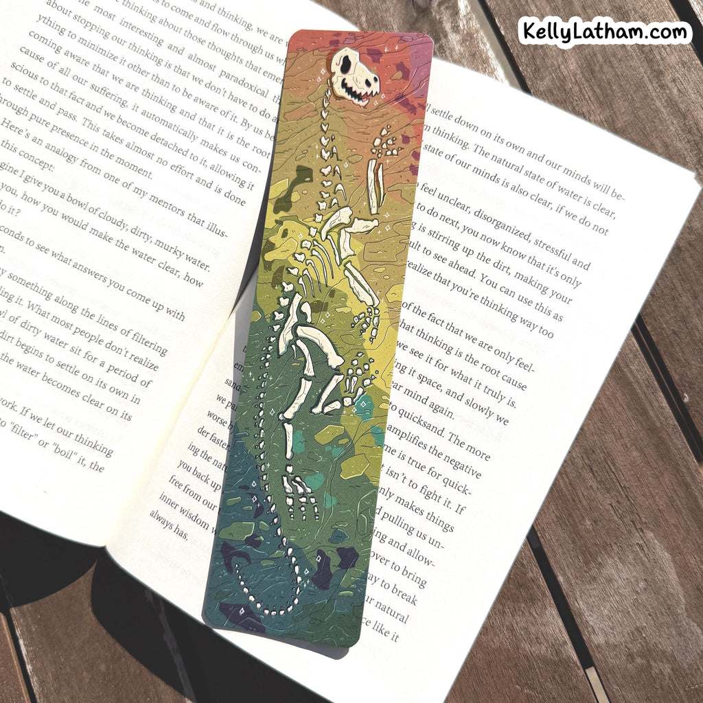 A long, narrow illustrated bookmark with a skeleton and floral design, placed on open book pages over a wooden surface. The bookmark has a gradient background transitioning from green to orange and purple, with a white skeleton motif running vertically.