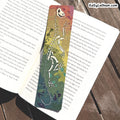 A long, narrow illustrated bookmark with a skeleton and floral design, placed on open book pages over a wooden surface. The bookmark has a gradient background transitioning from green to orange and purple, with a white skeleton motif running vertically.