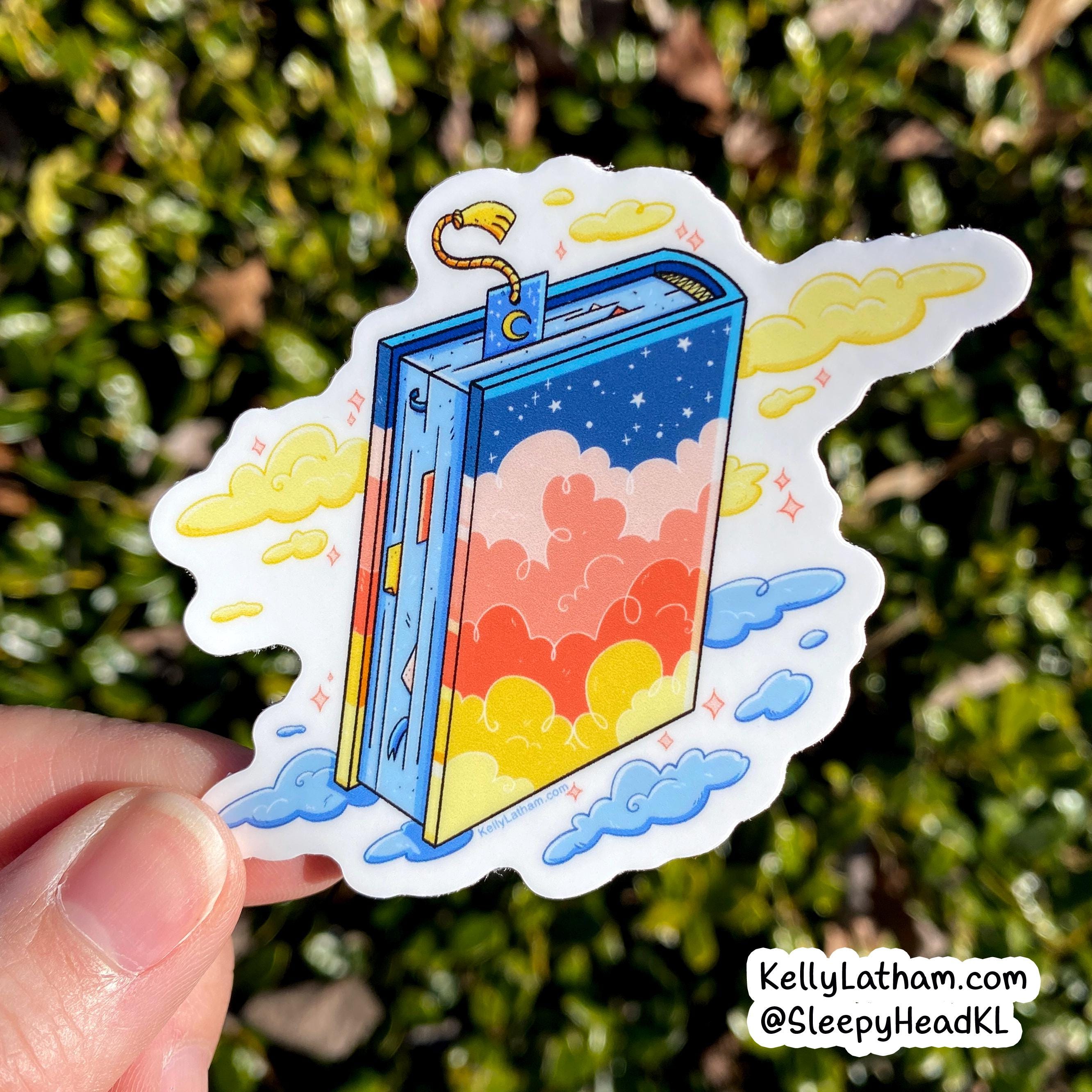 A hand holds a die-cut sticker featuring a whimsical open book with a blue starry sky cover and pastel clouds in yellow, pink, orange, and red. The background is blurred green foliage.
