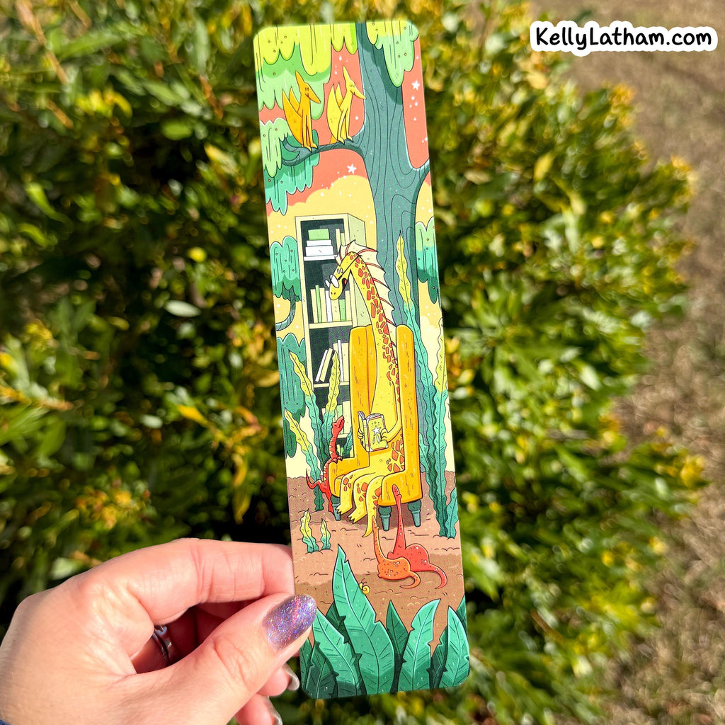 A hand holds a long, narrow illustrated bookmark featuring a dinosaur sitting on a yellow throne in front of a bookshelf, with a forest and house in the background. The hand is in front of blurred green foliage, and the text 'KellyLatham.