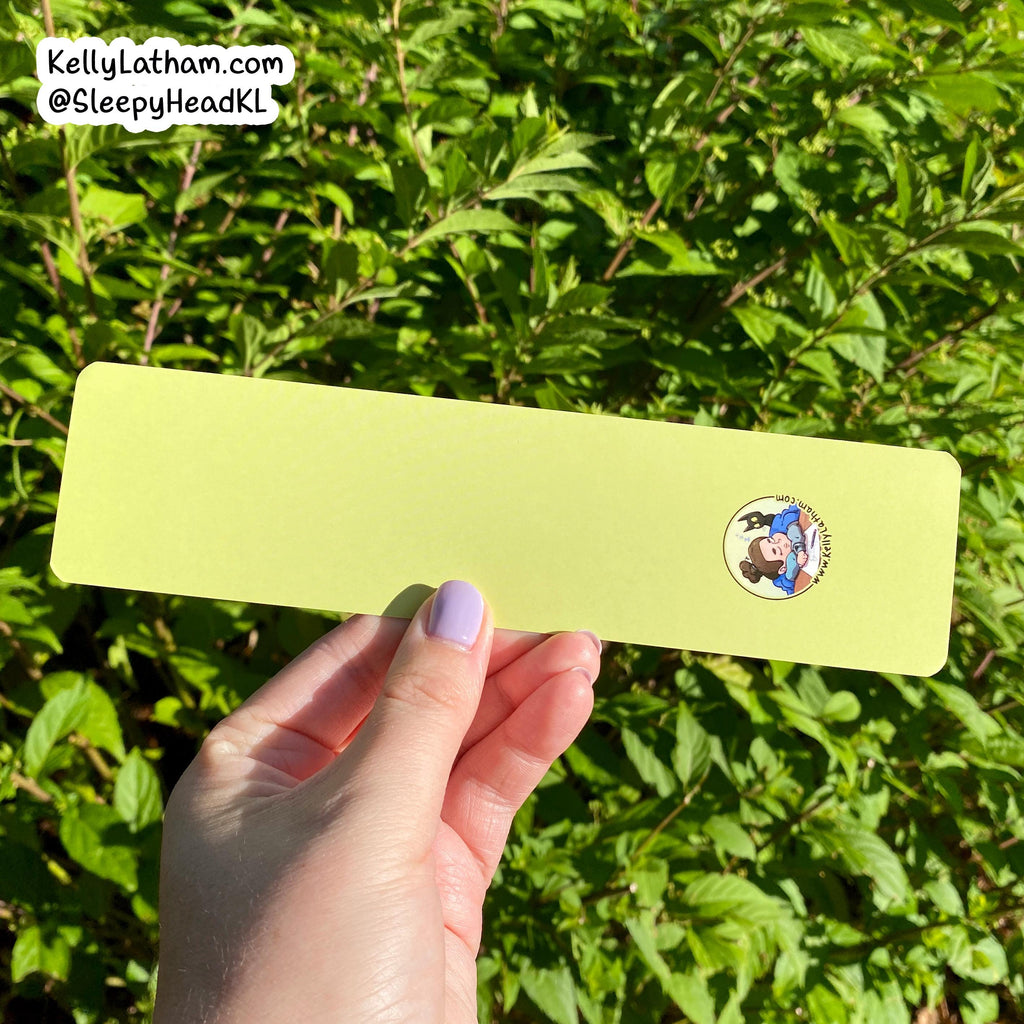 A hand with lavender nail polish holds a long, light yellow-green rectangular card or sticker against a background of green outdoor foliage. The card features a small circular cartoon illustration in the upper right corner.