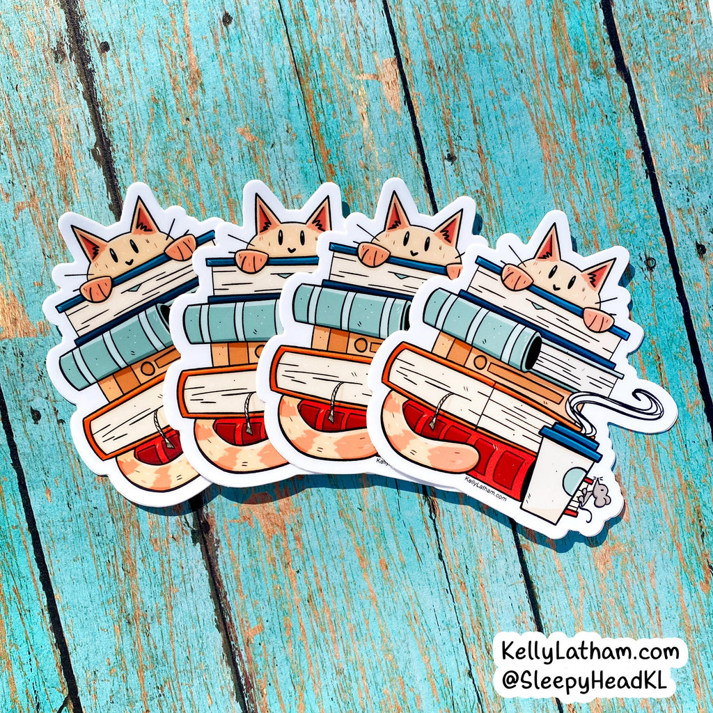 Four cartoon cat stickers stacked on books, arranged on a distressed turquoise wooden surface. The stickers show a cute illustrated cat peeking over a stack of books with a mug in the foreground.