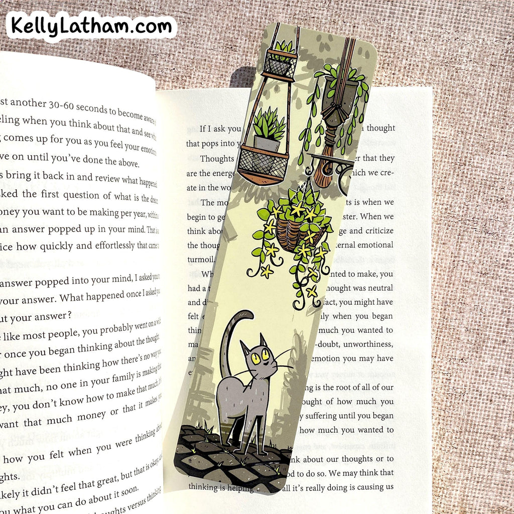 A decorative paper bookmark featuring an illustration of a gray cat sitting under hanging green plants, placed on an open book with printed text. The bookmark is photographed flat on a textured beige fabric background, with the website name KellyLatham.
