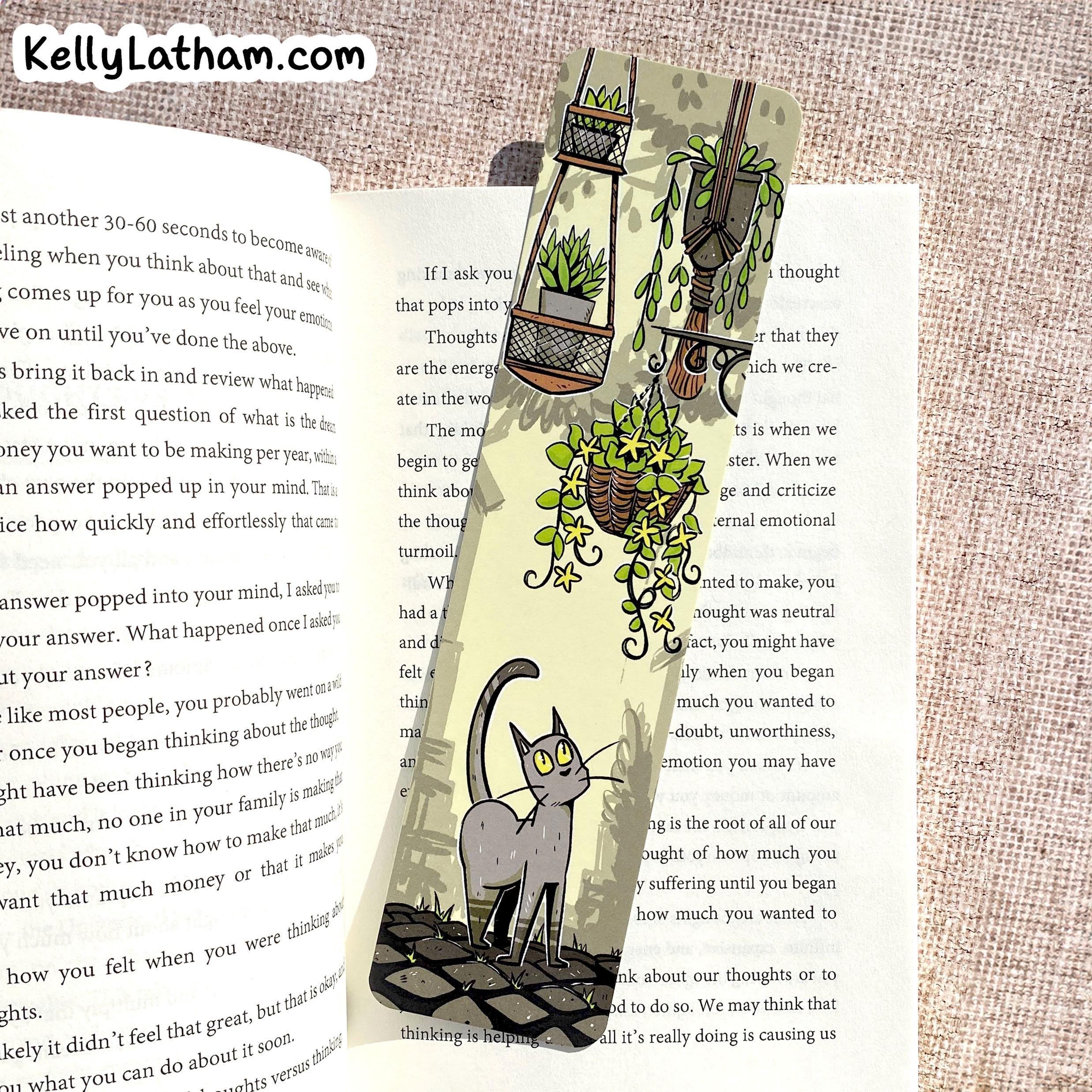 A decorative paper bookmark featuring an illustration of a gray cat sitting under hanging green plants, placed on an open book with printed text. The bookmark is photographed flat on a textured beige fabric background, with the website name KellyLatham.