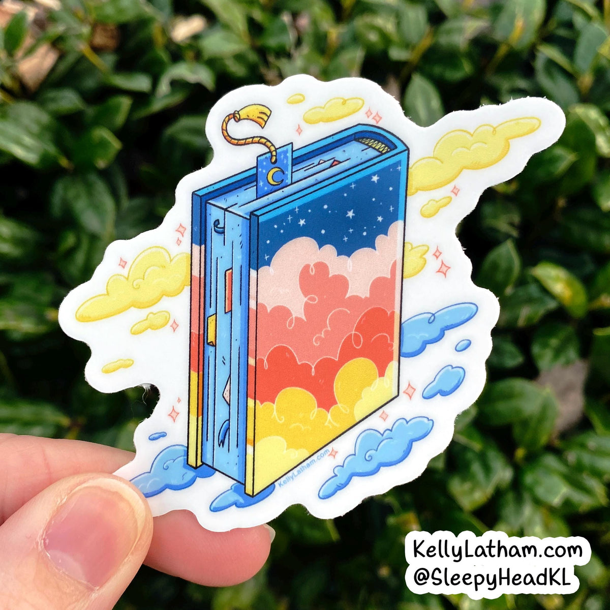 A hand holds a die-cut sticker featuring a whimsical illustrated book with a blue cover, yellow clouds, and pastel sky details, set against a blurred green leaf background.