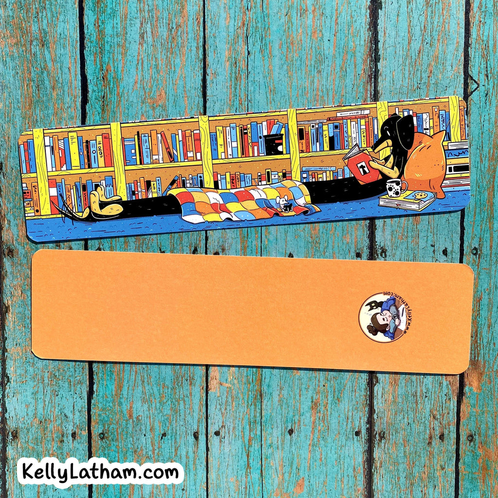 An illustrated book mark or bookmark featuring a cozy library scene with bookshelves, a person reading, and a colorful quilt, shown above a flat orange rectangular piece with a small circular logo and the text KellyLatham.com.