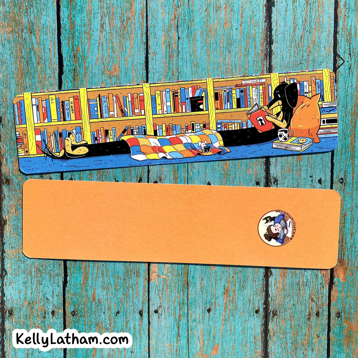 An illustrated book mark or bookmark featuring a cozy library scene with bookshelves, a person reading, and a colorful quilt, shown above a flat orange rectangular piece with a small circular logo and the text KellyLatham.com.