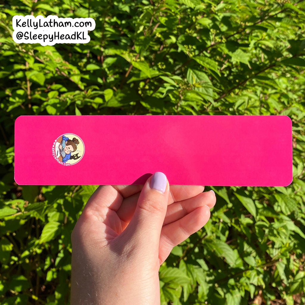A hand with lavender nail polish holds a bright pink rectangular sticker or card outdoors against a background of green leaves. The sticker has a small circular cartoon character graphic on the left side.