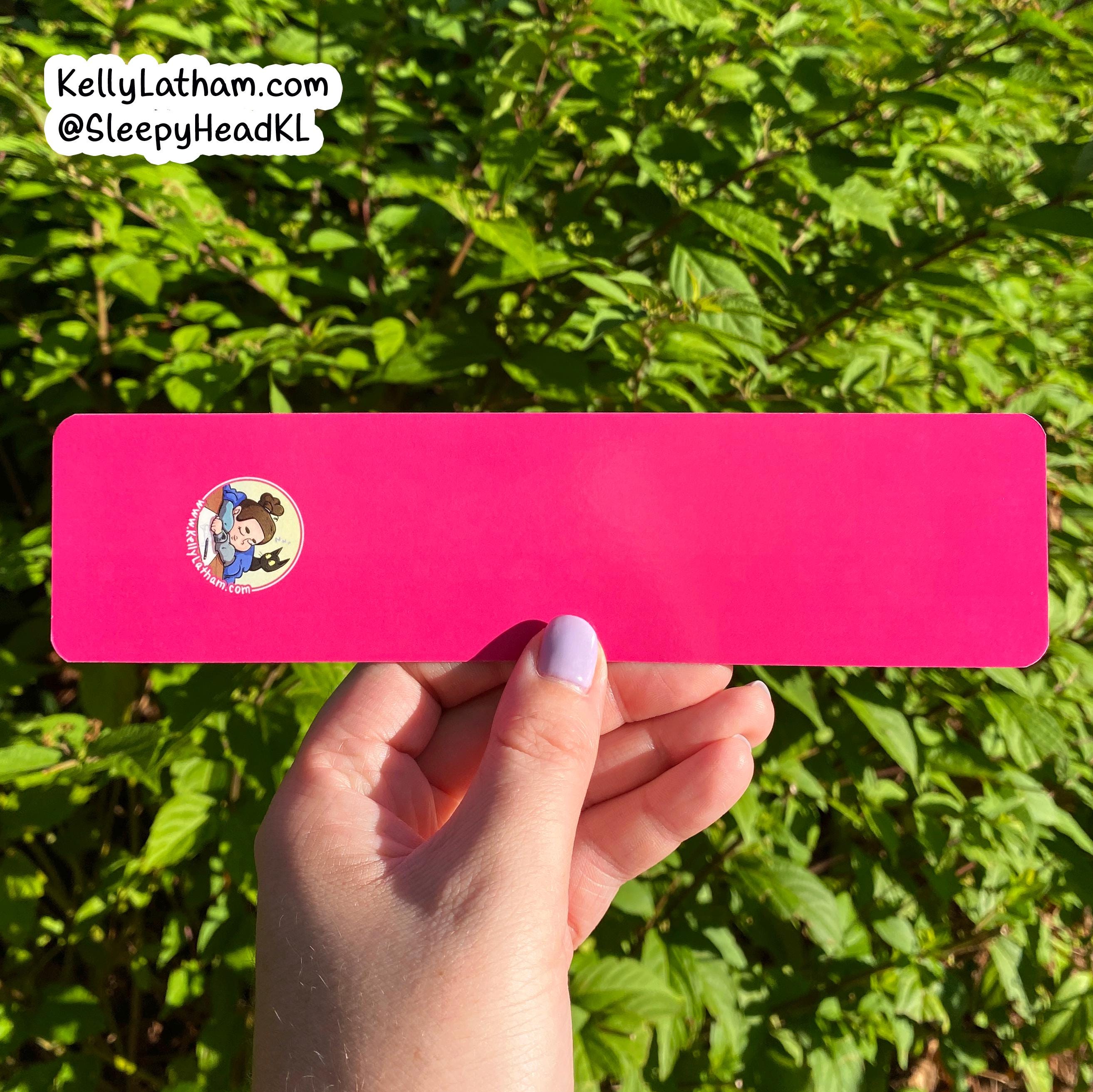 A hand with lavender nail polish holds a bright pink rectangular sticker or card outdoors against a background of green leaves. The sticker has a small circular cartoon character graphic on the left side.