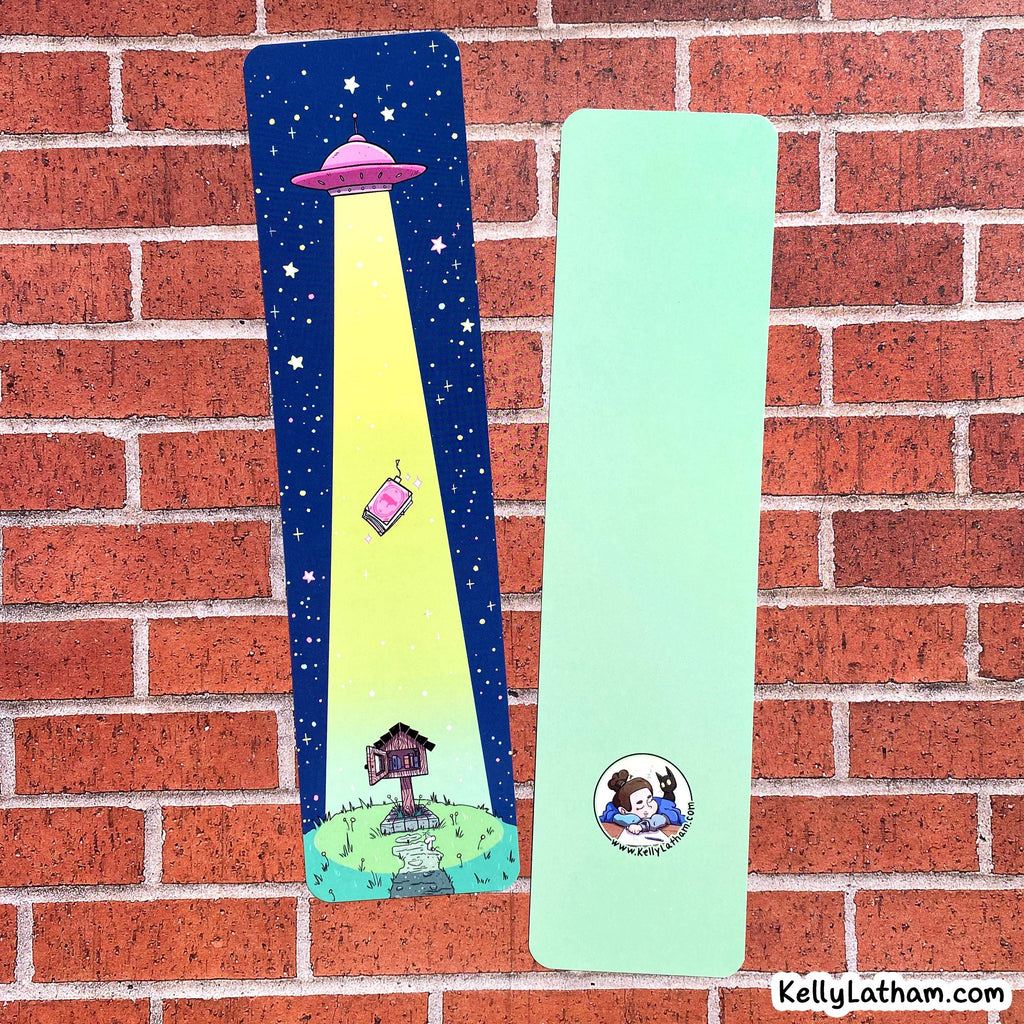 Two personalized bookmarks displayed on a red brick wall. One bookmark is dark blue with a yellow spotlight graphic of a pink house and a UFO, and the other is mint green with a custom cartoon illustration of a couple and the text 'Kelly & Liam - 2025'.