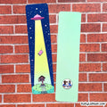 Two personalized bookmarks displayed on a red brick wall. One bookmark is dark blue with a yellow spotlight graphic of a pink house and a UFO, and the other is mint green with a custom cartoon illustration of a couple and the text 'Kelly & Liam - 2025'.