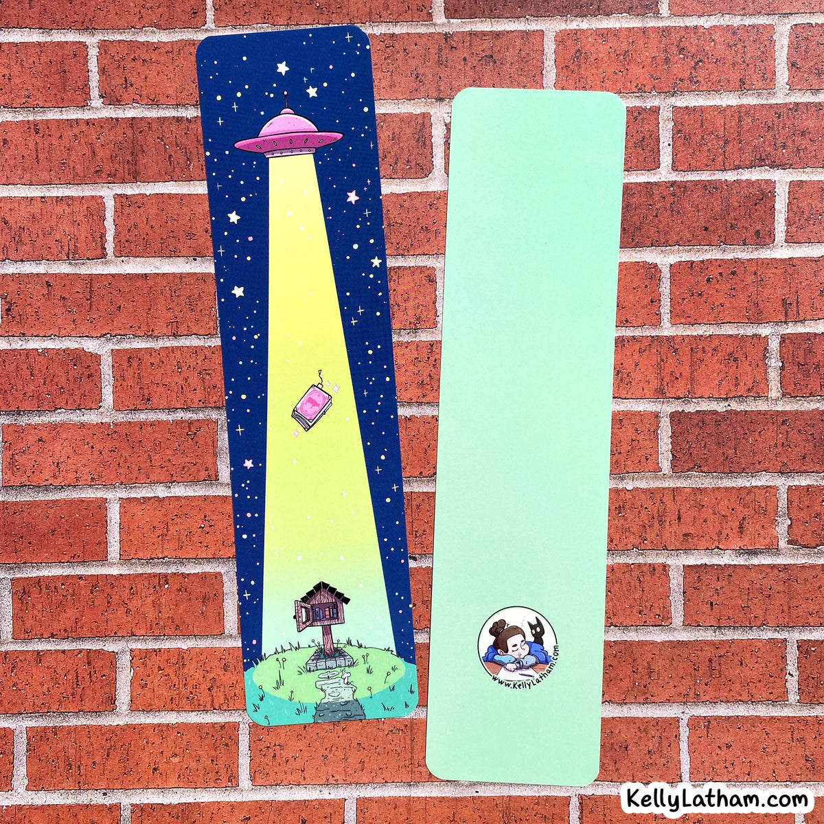 Two personalized bookmarks displayed on a red brick wall. One bookmark is dark blue with a yellow spotlight graphic of a pink house and a UFO, and the other is mint green with a custom cartoon illustration of a couple and the text 'Kelly & Liam - 2025'.