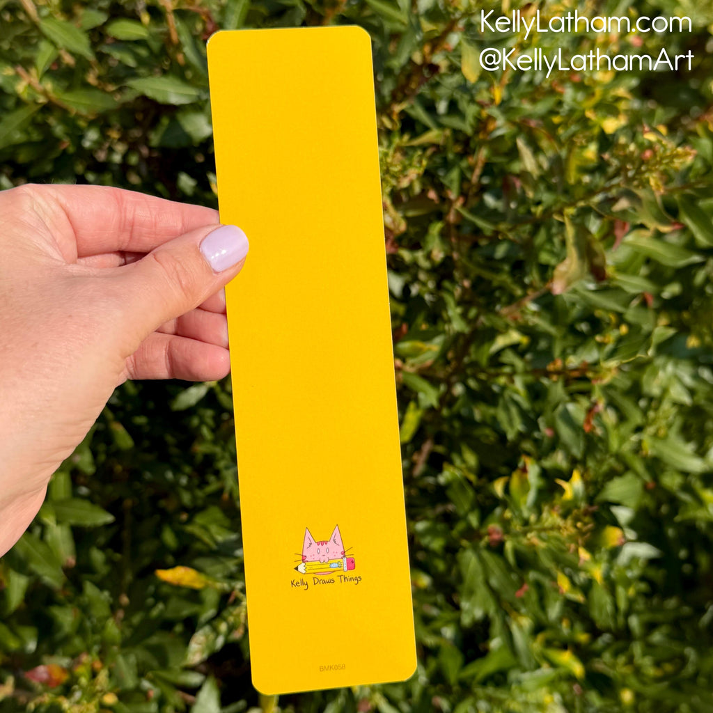 A hand holds a bright yellow rectangular art print or card featuring a small pink cat illustration and the text 'Kitty Daze Thing' against a background of green foliage. A watermark in the top right reads 'KellyLatham.