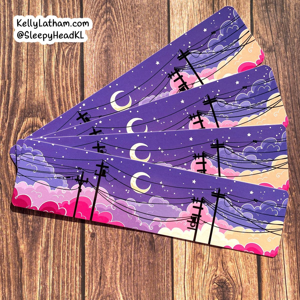 Four rectangular coasters with a whimsical night sky design, fanned out on a brown wooden surface. The design features a purple sky with crescent moons, stars, pink clouds, and black power lines.