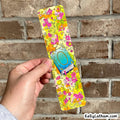 A hand holds a long, narrow bookmark or card featuring a bright yellow background with a dense floral pattern in pink, orange, green, and white, along with a small illustrated book graphic. The background is a real brick wall.