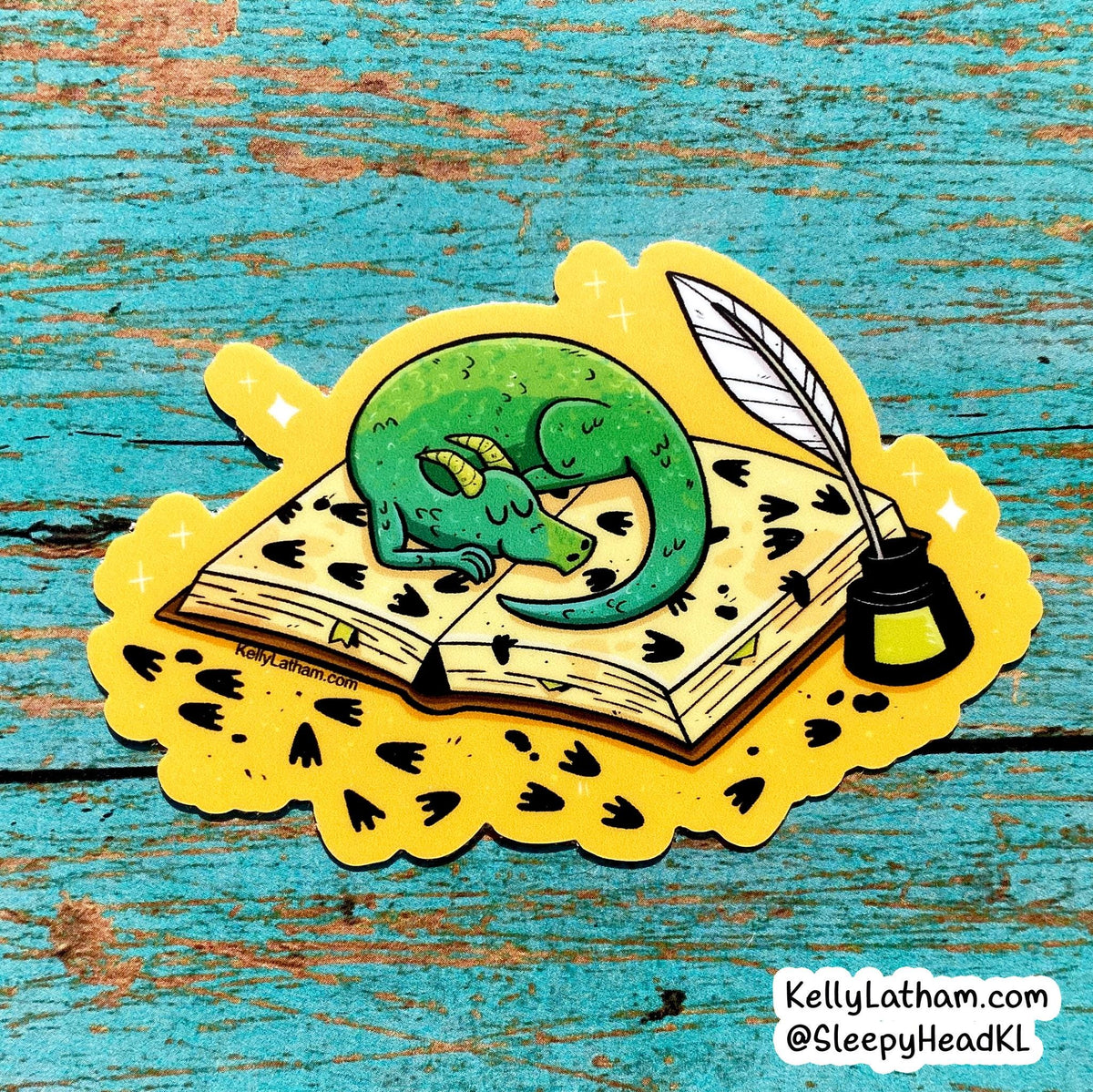 A die-cut sticker featuring a green sleeping dragon curled over an open book with black heart prints, placed on a yellow scalloped surface with a quill and ink bottle. The sticker is photographed on a distressed turquoise wooden surface, with a small KellyLatham.
