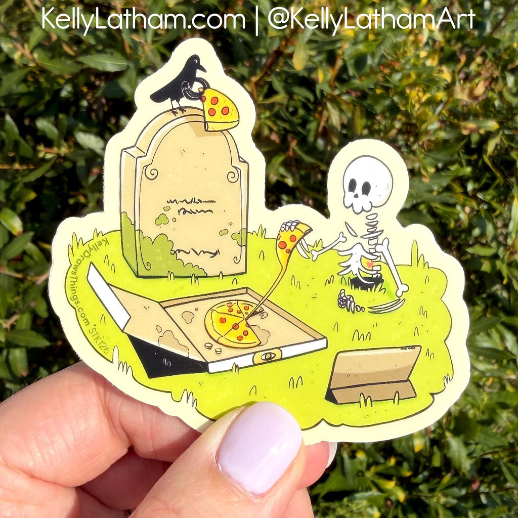 A die-cut sticker featuring a cartoon skeleton eating pizza next to a tombstone with a crow, held by a hand against a green hedge background. The sticker has a cream border and is shown outdoors with the Kelly Latham branding visible at the top.