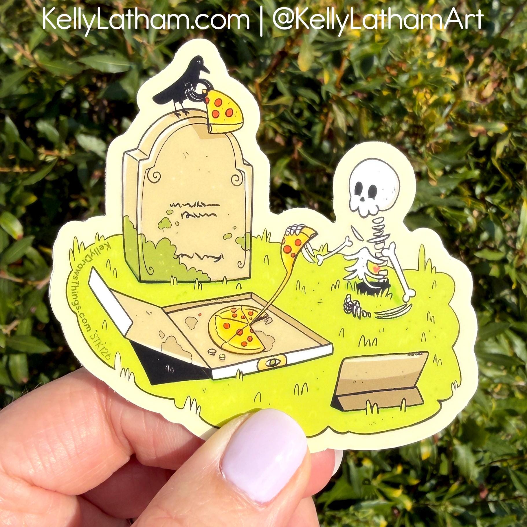 A die-cut sticker featuring a cartoon skeleton eating pizza next to a tombstone with a crow, held by a hand against a green hedge background. The sticker has a cream border and is shown outdoors with the Kelly Latham branding visible at the top.