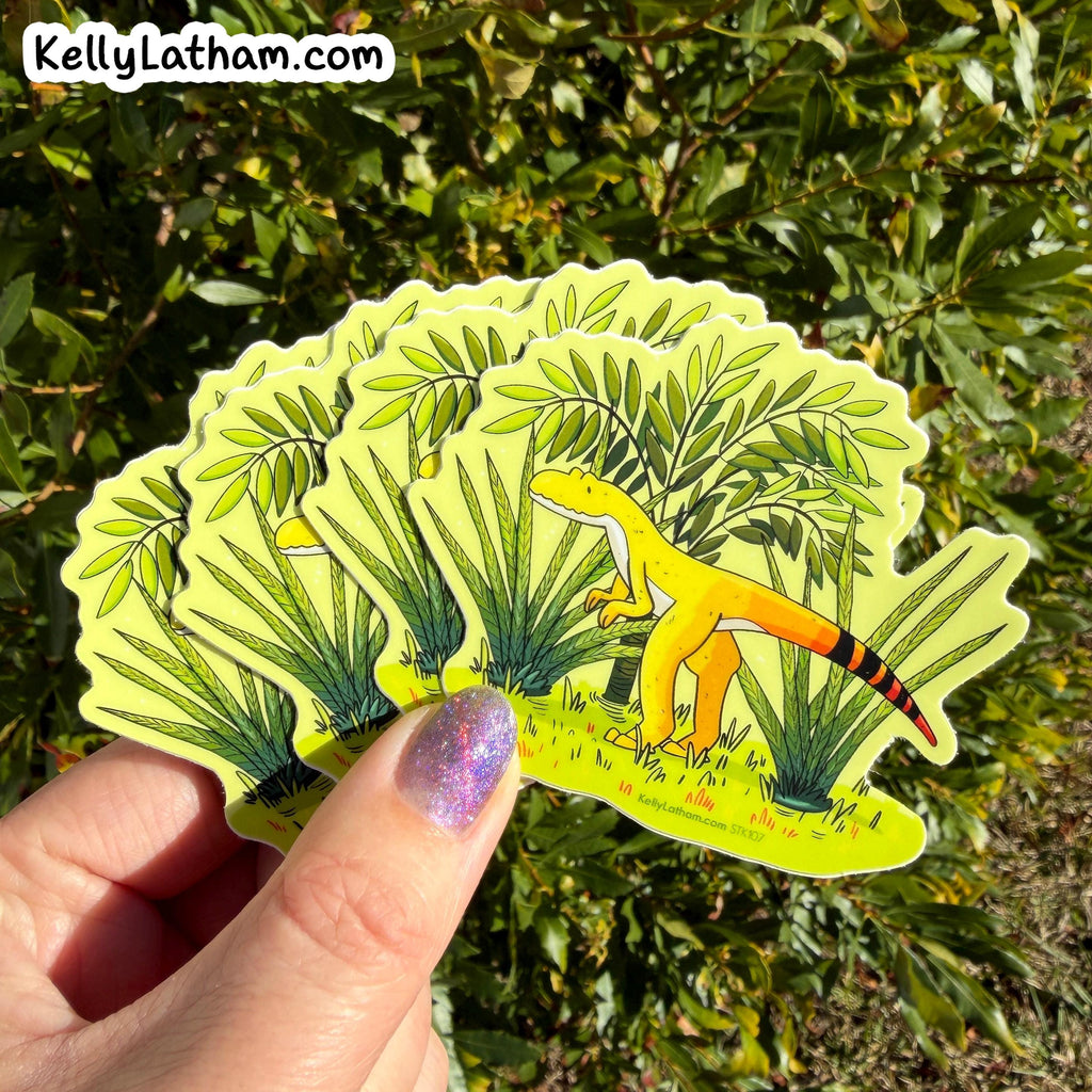 A hand holds a fan of die-cut stickers featuring a yellow cartoon dinosaur among green plants, photographed outdoors in front of green foliage. The top-left corner shows the text KellyLatham.