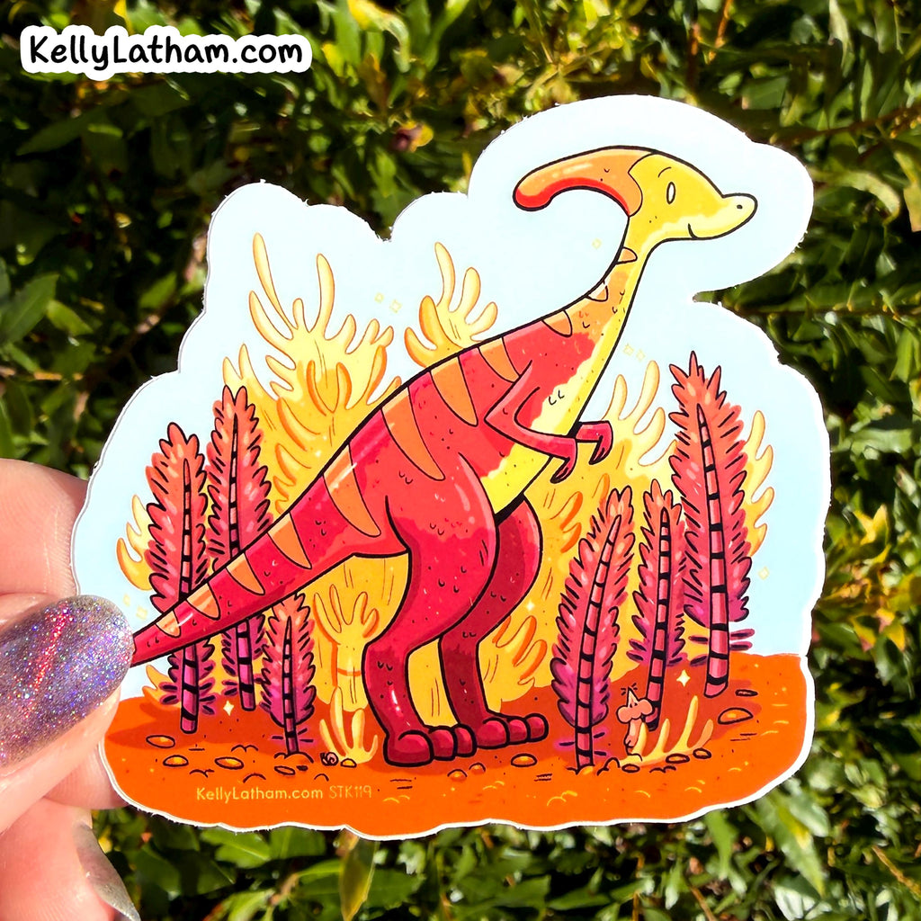 A die-cut sticker featuring a cartoon red and yellow dinosaur standing in front of orange flames, held by a hand against a background of green foliage. The sticker has a white border and the brand name 'KellyLatham.