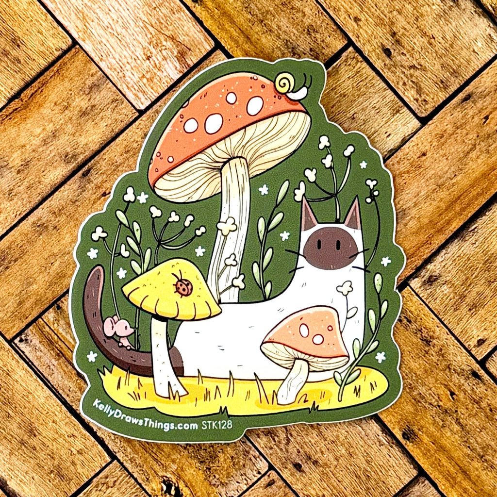 A die-cut sticker featuring a whimsical illustration of a white cat with a dark face among mushrooms and small white flowers, placed on a wooden plank background. The sticker shows the text 'KellyDrewsThings.