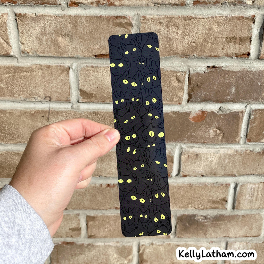 A hand holds a slim black bookmark or card featuring a repeating pattern of black spiders with glowing yellow eyes, set against a rustic brick wall background.