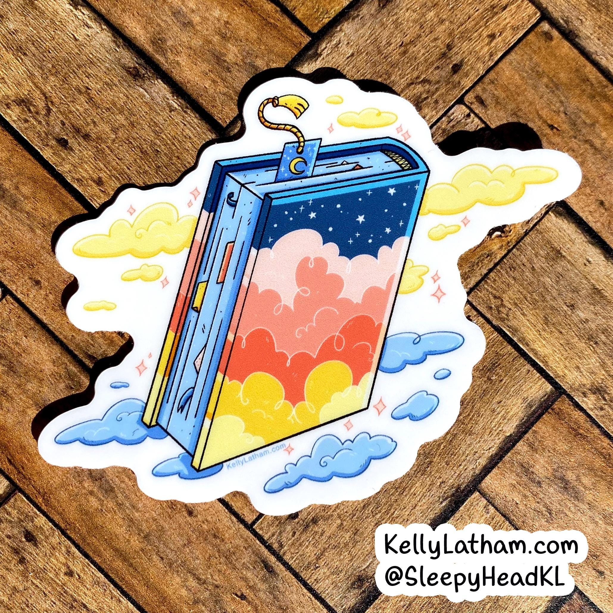 A die-cut sticker featuring a colorful illustrated book with a starry blue sky cover, surrounded by yellow clouds, placed on a brown wooden surface. A watermark in the bottom right reads KellyLatham.