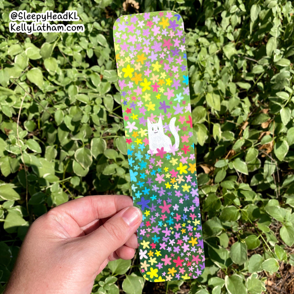 A hand holds a long rectangular bookmark or card covered in a colorful floral pattern and a white cat illustration, set against a background of green outdoor leaves. The watermark in the top left reads @SleepyHeadKL KellyLatham.
