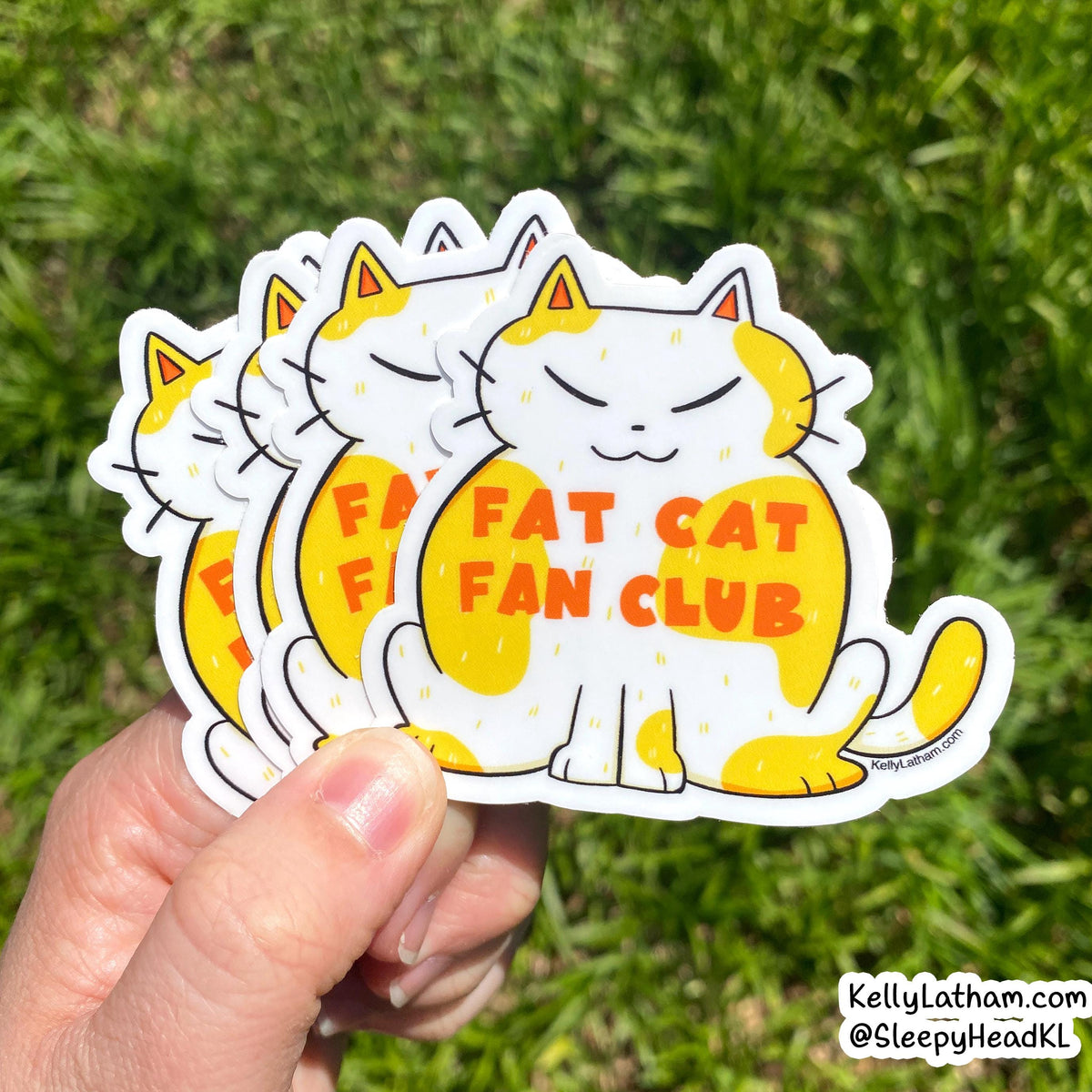 A hand holds a stack of die-cut stickers featuring a cartoon white cat with yellow patches and the text 'FAT CAT FAN CLUB' in orange, against a blurred green grass background.